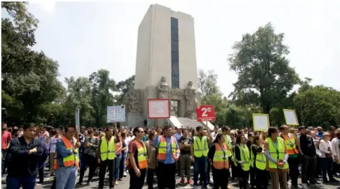 Fotografía de una concentración comunitaria frente a un monumento, con personas usando chalecos reflectantes y carteles.