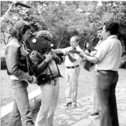 Fotografía en blanco y negro de un equipo de filmación: una persona sostiene una cámara de video mientras otras colaboran en exteriores.