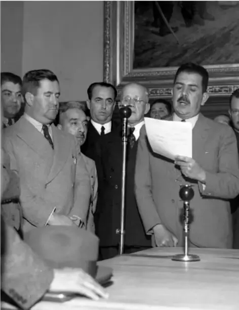 Fotografía en blanco y negro del presidente Lázaro Cárdenas del Río leyendo un documento ante un micrófono, rodeado de funcionarios y personas en un salón oficial.