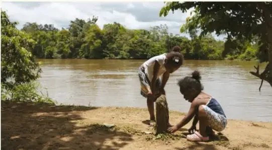 Fotografía de una mujer y una niña reunidas en la orilla de un río con vegetación, posiblemente realizando una actividad cotidiana en una comunidad.