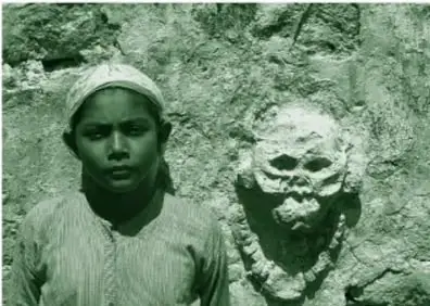 Fotografía en blanco y negro (impresa en tinta verde): un niño con paliacate en la cabeza recargado en una pared de piedra junto a un relieve de calavera con antifaz y un collar tallado.