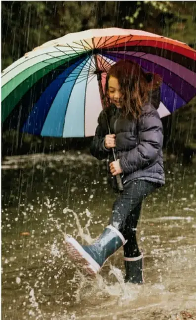 Niña con paraguas de colores saltando en un charco bajo la lluvia.