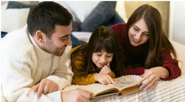 Fotografía de una familia (papá, mamá e hija pequeña) recostada junta, sonriendo y leyendo un libro abierto.