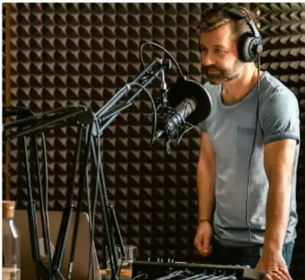 Fotografía de un hombre con audífonos frente a un micrófono en un estudio de grabación con paneles acústicos, ilustrando el trabajo de sonido y doblaje.