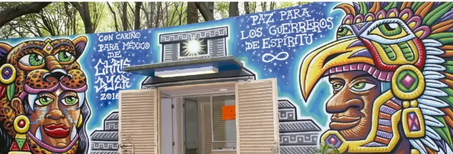 Mural de colores vibrantes en una pared exterior de una construcción, con figuras de un felino (jaguar) antropomorfe a la izquierda y un guerrero emplumado (guerrero azteca) a la derecha; incluye textos "Con cariño para México de Chi'as... 2018" y "Paz para los guerreros de espíritu" con un símbolo de infinito.