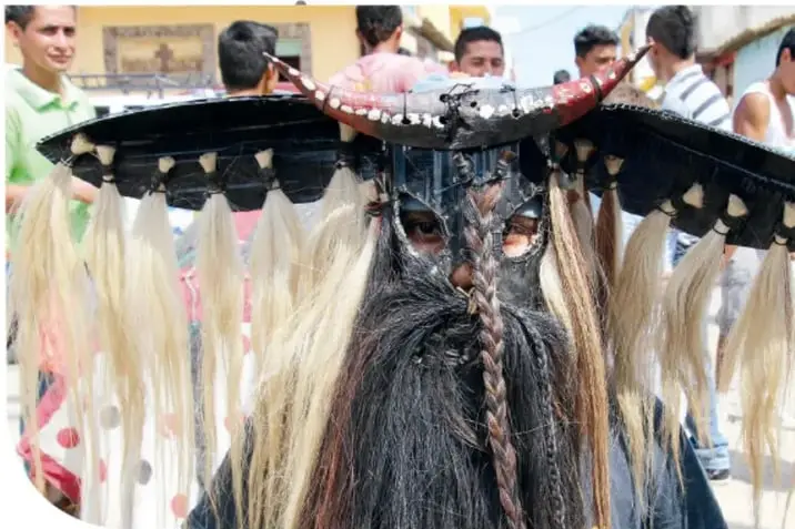 Grupo de personas sentadas, algunas con máscaras de diablo y vestimenta tradicional, una de ellas sostiene un instrumento.