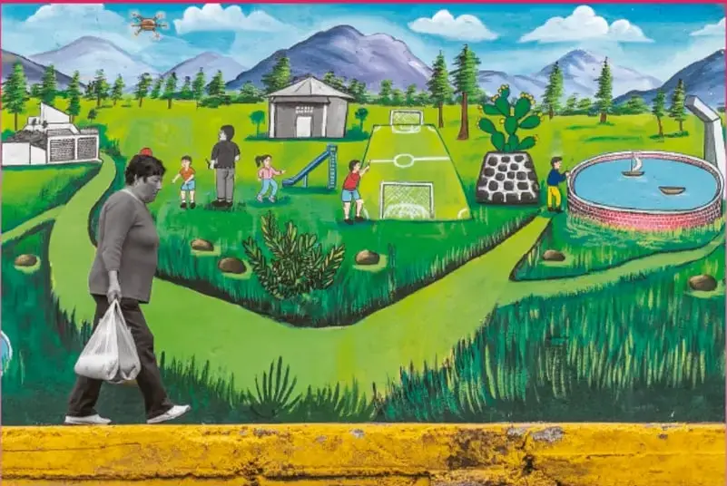Fotografía de una persona caminando por una banqueta amarilla con bolsa en la mano; al fondo un mural pintado con un parque, cancha de fútbol, tobogán, lámina de agua, niñas y niños jugando, árboles y montañas.