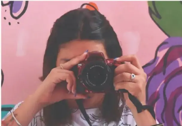 Fotografía de una estudiante (Gimenna Schiaffini Rosales, Puebla) sosteniendo una cámara roja para tomar una fotografía, frente a un muro con arte urbano de colores.