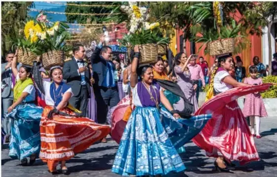 Fotografía de un desfile con personas vestidas con trajes tradicionales de distintas regiones de México, portando canastas y faldas de colores, en un contexto festivo.