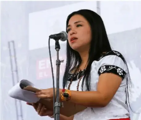 Fotografía de una mujer joven de rasgos indígenas, con cabello largo oscuro y camisa blanca bordada, hablando frente a un micrófono y sosteniendo un papel, durante una asamblea o evento comunitario.