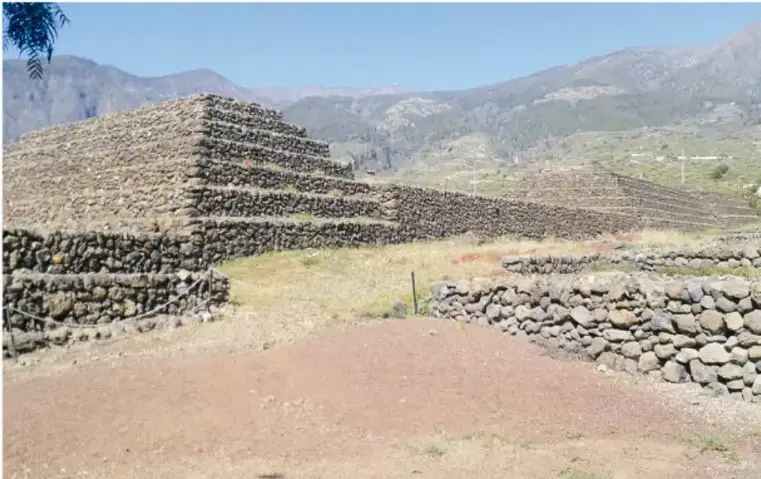 Fotografía de una pirámide escalonada de Güímar construida con piedras volcánicas, con un camino de tierra al frente y montañas al fondo.