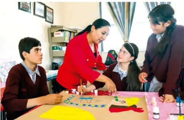 Docente en chamarra roja trabajando con tres estudiantes sobre un cartel con materiales de colores en el aula.