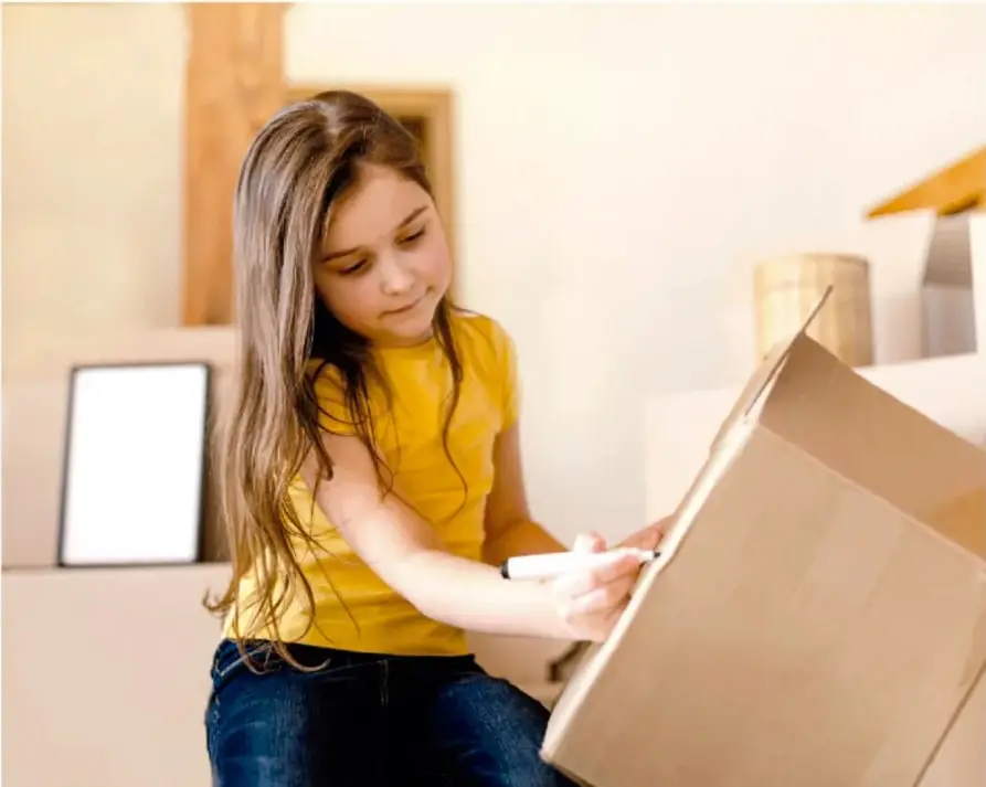 Fotografía de una niña con playera amarilla que escribe o dibuja sobre una caja de cartón grande, con una televisión de cartón al fondo.
