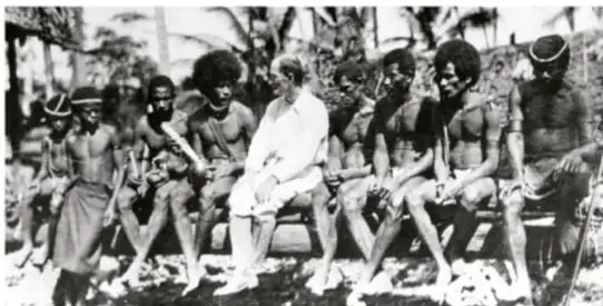 Fotografía en blanco y negro de un grupo de personas (comunidad indígena o africana) sentadas junto a un hombre de traje, probablemente un investigador, en un entorno comunitario.