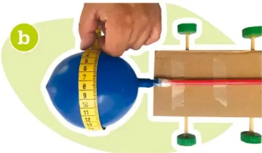 Fotografía de un globo azul inflado a menor capacidad, con cinta métrica midiendo su circunferencia, colocado en el mismo dispositivo de madera. Se marca con la letra b.