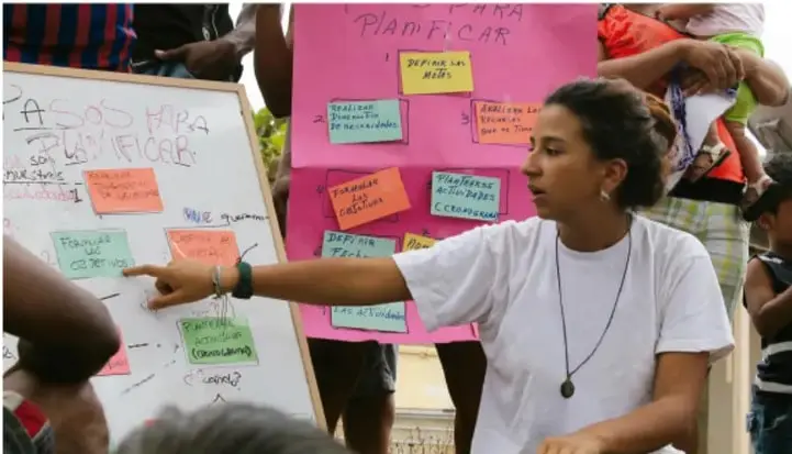Fotografía de una facilitadora comunitaria explicando frente a cartulinas con notas adhesivas los 'Pasos para planificar' a un grupo de personas, en un taller comunitario al aire libre.