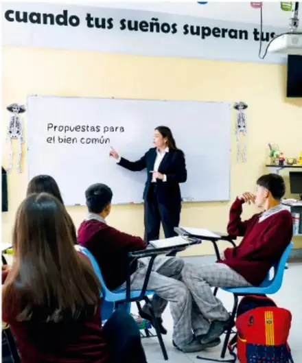 Fotografía de un aula de telesecundaria: la maestra frente al pizarrón que dice 'Propuestas para el bien común', con bancas en círculo y estudiantes participando; en la pared se lee 'cuando tus sueños superan tus...'