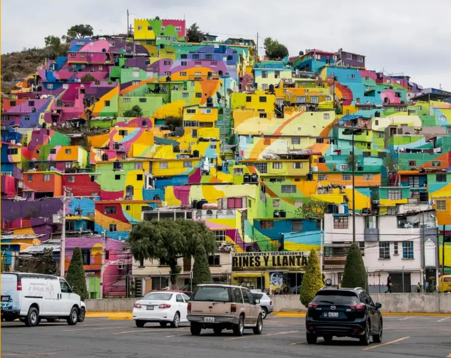 Fotografía de un barrio urbano en pendiente con casas pintadas de múltiples colores, con un estacionamiento y vehículos al frente; rótulo 'COMPRA VENTA Y REPARACIÓN DE RINES Y LLANTAS'.