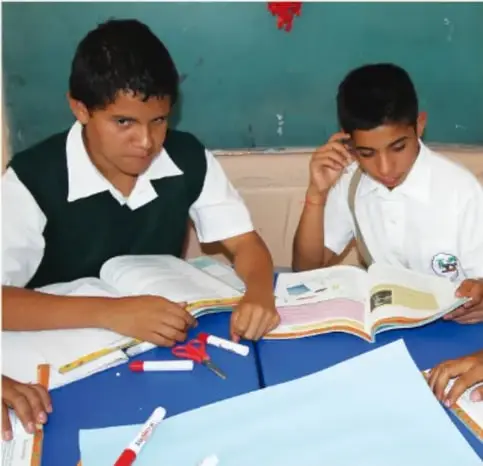 Fotografía de dos estudiantes de secundaria con uniforme trabajando juntos en una mesa con cuaderno, colores y papel, realizando el boceto de su estructura.