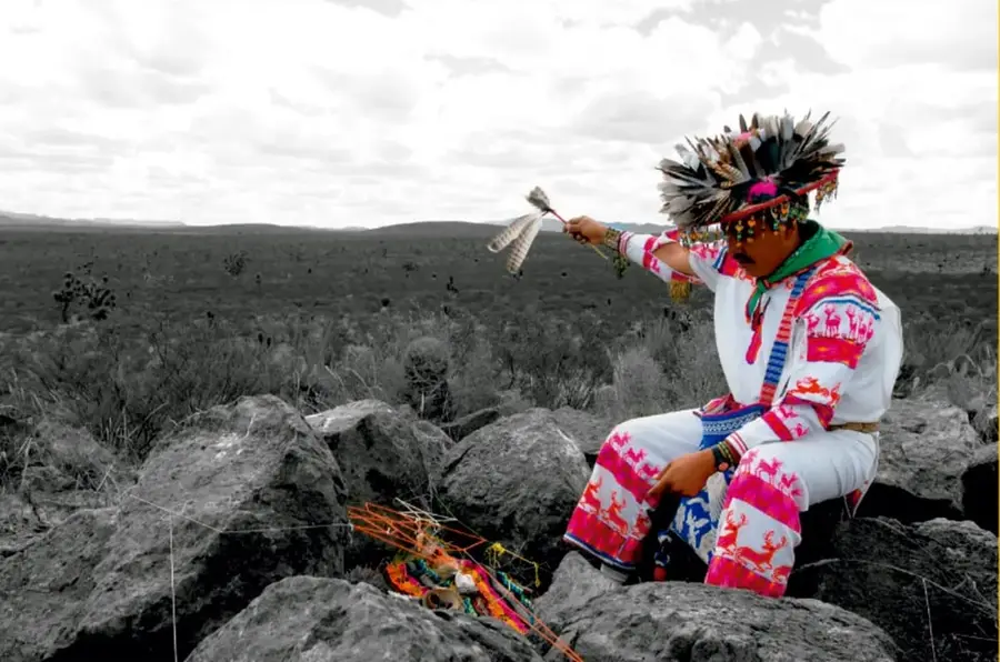 Fotografía en blanco y negro con detalles a color de un joven con traje tradicional emplumado, sentado sobre rocas en un paisaje desértico con nubes; pie de foto: 'Salatiel Barragán Santos, VERACRUZ'.