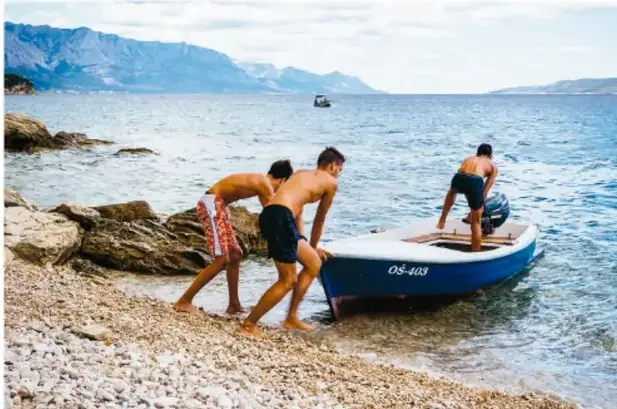 Fotografía de tres jóvenes empujando una lancha hacia el mar sobre un suelo pedregoso. Se observa el esfuerzo por vencer la fricción estática.