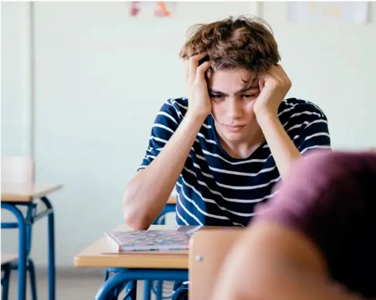 Fotografía de un adolescente sentado en su pupitre en el aula, con la cabeza entre las manos y expresión preocupada o abrumada; al frente se ve el hombro de otro compañero.
