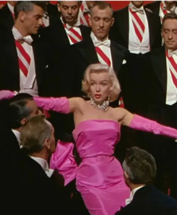 Fotografía a color de Marilyn Monroe con vestido rosa y guantes largos, rodeada de hombres con esmoquin y corbata roja (escena de la película "Los que no besan a su vecino" o similar).