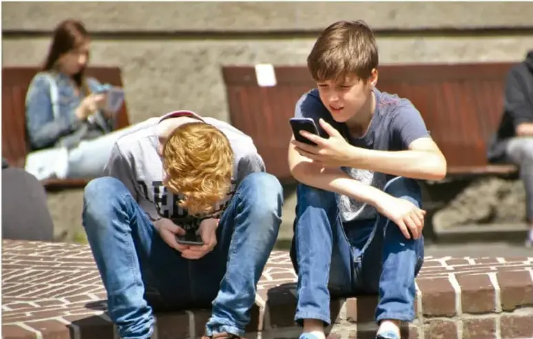 Fotografía de dos niños sentados en el suelo usando teléfonos móviles, con una persona al fondo también con un dispositivo. Ilustra el tema del uso de dispositivos móviles en la infancia.