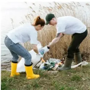 Fotografía de dos personas con botas amarillas y guantes recogiendo basura y desechos en un área natural junto a un cuerpo de agua, como ejemplo de acción comunitaria para cuidar el ambiente.