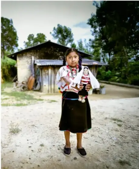 Fotografía de una niña indígena de pie frente a una casa rural de madera, con árboles al fondo. Viste blusa bordada, falda oscura y sostiene un muñeco de trapo. Representa la diversidad cultural y la identidad personal.