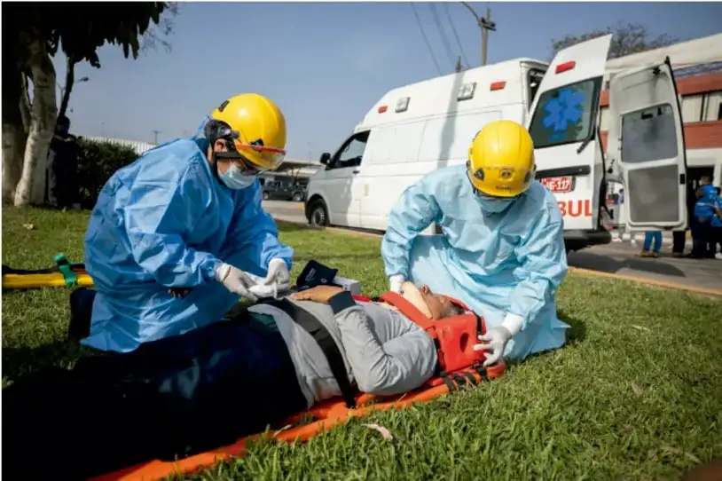 Fotografía de dos paramédicos con equipo de protección (overoles azules, cascos amarillos, guantes) atendiendo a una persona herida recostada en una camilla naranja sobre el pasto. Al fondo, una ambulancia blanca con puertas abiertas y personal de emergencia.