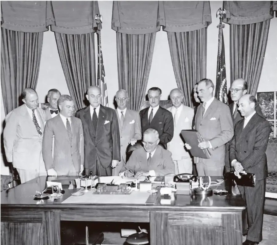 Fotografía en blanco y negro de una reunión diplomática. Varios hombres con trajes formales están de pie alrededor de un escritorio; uno de ellos, sentado, firma un documento. Al fondo hay cortinas y banderas. La imagen ilustra la firma de un acuerdo internacional, probablemente relacionado con el bloque socialista.