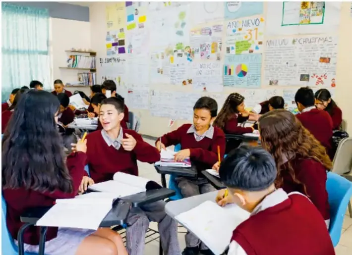 Fotografía de estudiantes de secundaria trabajando en equipo en el aula, con murales y trabajos en las paredes; unos escriben y otros conversan.