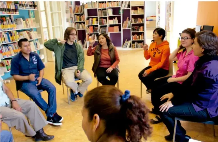 Fotografía de un grupo de estudiantes y una persona adulta sentados en círculo dentro de una biblioteca, conversando durante una asamblea de aula.