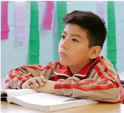 Fotografía de un niño con sudadera roja y beige recargado sobre un libro abierto, mirando hacia un lado en un salón decorado con listones de papel rosa y verde.
