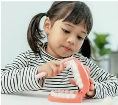 Niña de primaria con camisa a rayas observando y señalando un modelo grande de dientes, representando el oficio del odontólogo.