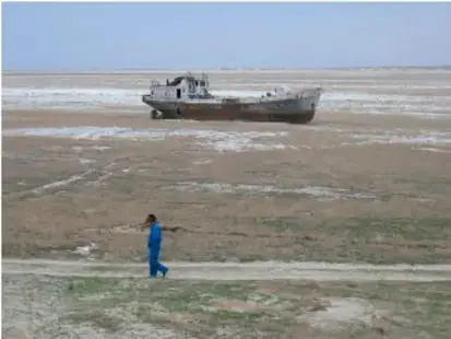 Fotografía de un barco varado sobre un lecho seco de un cuerpo de agua reducido por sequía, con una persona caminando en primer plano; ilustra los efectos del cambio climático.