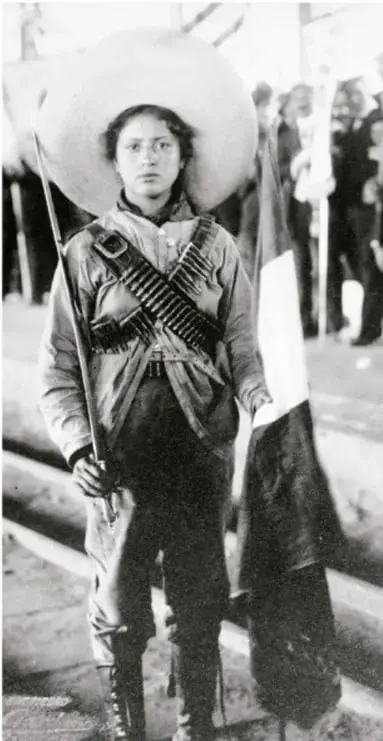 Fotografía en blanco y negro de una mujer revolucionaria con cananas cruzadas al pecho, sosteniendo un rifle y una bandera, con un sombrero grande; representa la participación femenina en la Revolución Mexicana.