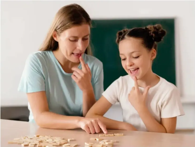 Fotografía de una maestra y una estudiante sentadas ante una mesa con fichas de letras; la maestra señala su boca y la niña imita el gesto, en un aula con pizarrón verde al fondo.