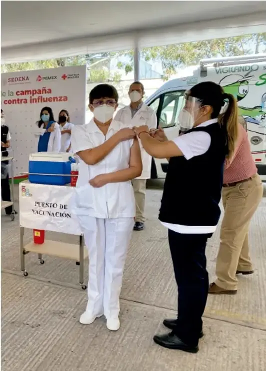 Fotografía de una campaña contra influenza: puesto de vacunación con personal de salud aplicando la vacuna a una usuaria; fondos con carteles institucionales y un vehículo de vigilancia.