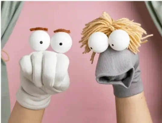 Dos títeres de guante (uno blanco con ojos grandes, otro gris con cabello amarillo) frente a un fondo rosa con cortinas verdes, ilustrando la creación de personajes con materiales diversos.