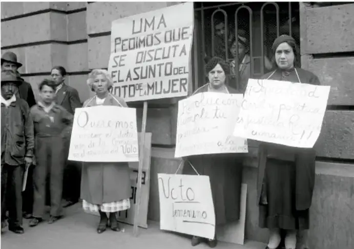 Fotografía histórica en blanco y negro de un grupo de mujeres sufragistas frente a un edificio, sosteniendo carteles con leyendas como 'UMA', 'Voto femenino' y 'PEDIMOS QUE SE DISCUTA EL ASUNTO DEL VOTO DE MUJER'.