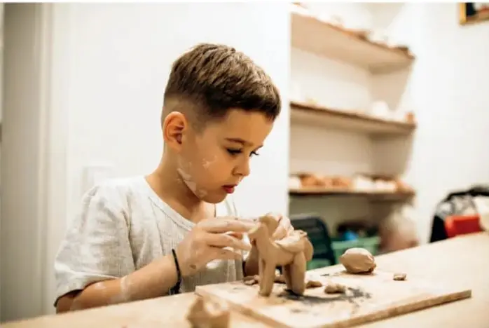 Fotografía de un niño con playera clara que modela una figura de arcilla sobre una tabla de madera; al fondo se ven estantes con materiales.