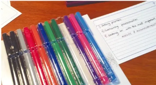 Fotografía de plumas de colores alineadas sobre un escritorio junto a una hoja de notas con ideas escritas del proyecto (Setting priorities, Gathering information, Working on it with the right people, Assess & accomplish).