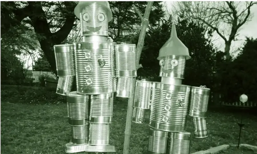 Fotografía de dos figuras humanas tipo 'hombre de hojalata' construidas con latas y envases metálicos reciclados de distintos tamaños, con detalles como tapas y piezas de metal; están en un jardín con árboles y pasto, apoyadas sobre una base clara.