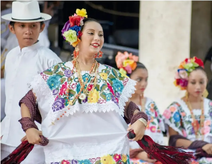 Fotografía de un grupo de danzantes con vestimenta tradicional: un joven con sombrero blanco y camisa blanca, y mujeres con blusas bordadas de colores vivos, collares y tocados florales, sosteniendo listones rojos. Representa las prácticas culturales de la comunidad.