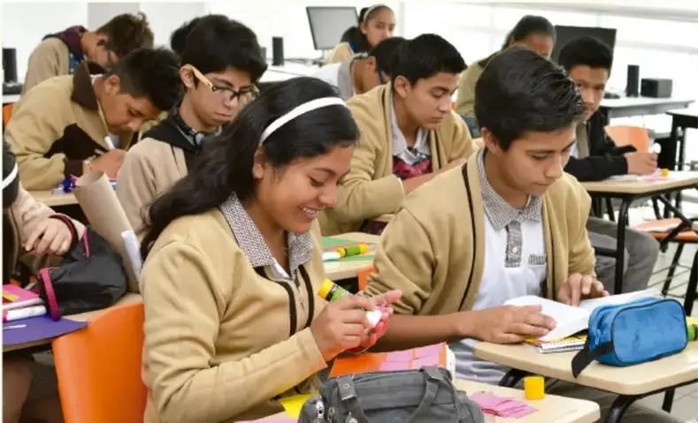 Fotografía de estudiantes de secundaria trabajando en equipo en el salón, tomando notas y sonriendo.