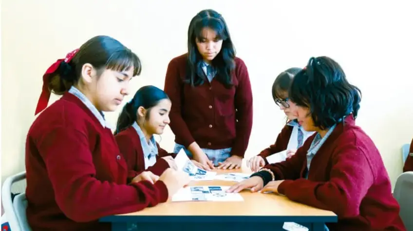 Fotografía de cinco estudiantes de telesecundaria con uniforme chamarra roja, reunidos alrededor de una mesa trabajando juntos sobre documentos y materiales del proyecto.