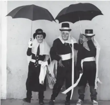 Fotografía en blanco y negro de tres danzantes con sombrillas negras, sombreros de ala y listones, posando frente a un muro; Danza de los paragüeros, 1991, Contla de Juan Cuamatzi, Tlaxcala, de Mariana Yampolsky (1925-2002).