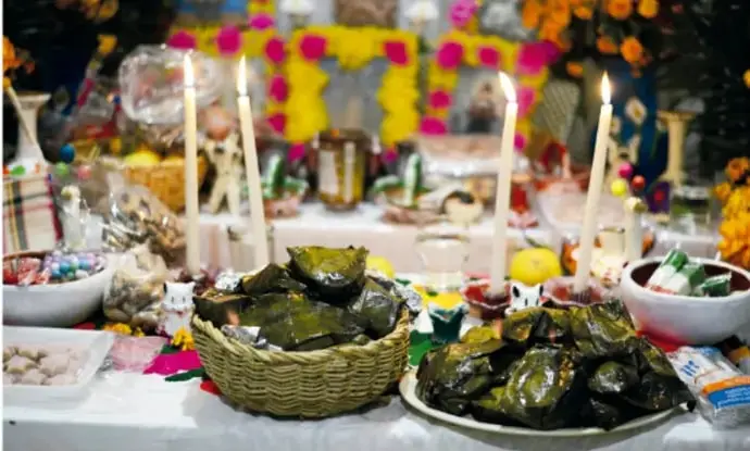 Altar de ofrendas con veladoras encendidas, canastas de tamales envueltos en hoja, flores, frutas y figuras ornamentales de colores.